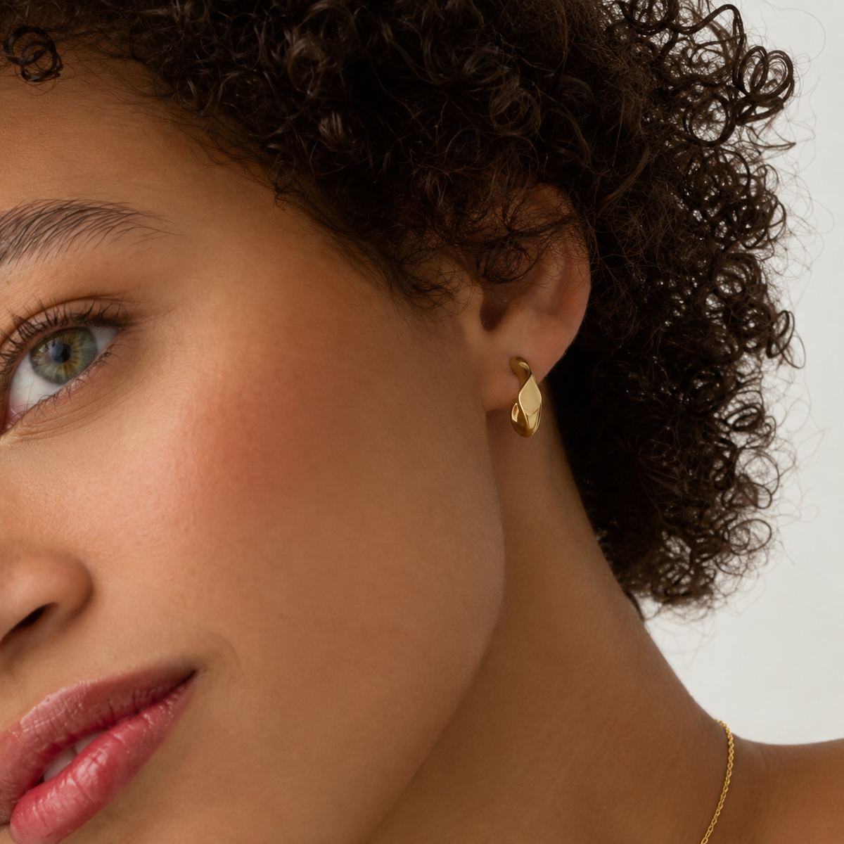 A close-up of a model with curly dark hair wearing a small gold hoop earring and a delicate gold necklace against a light neutral background.