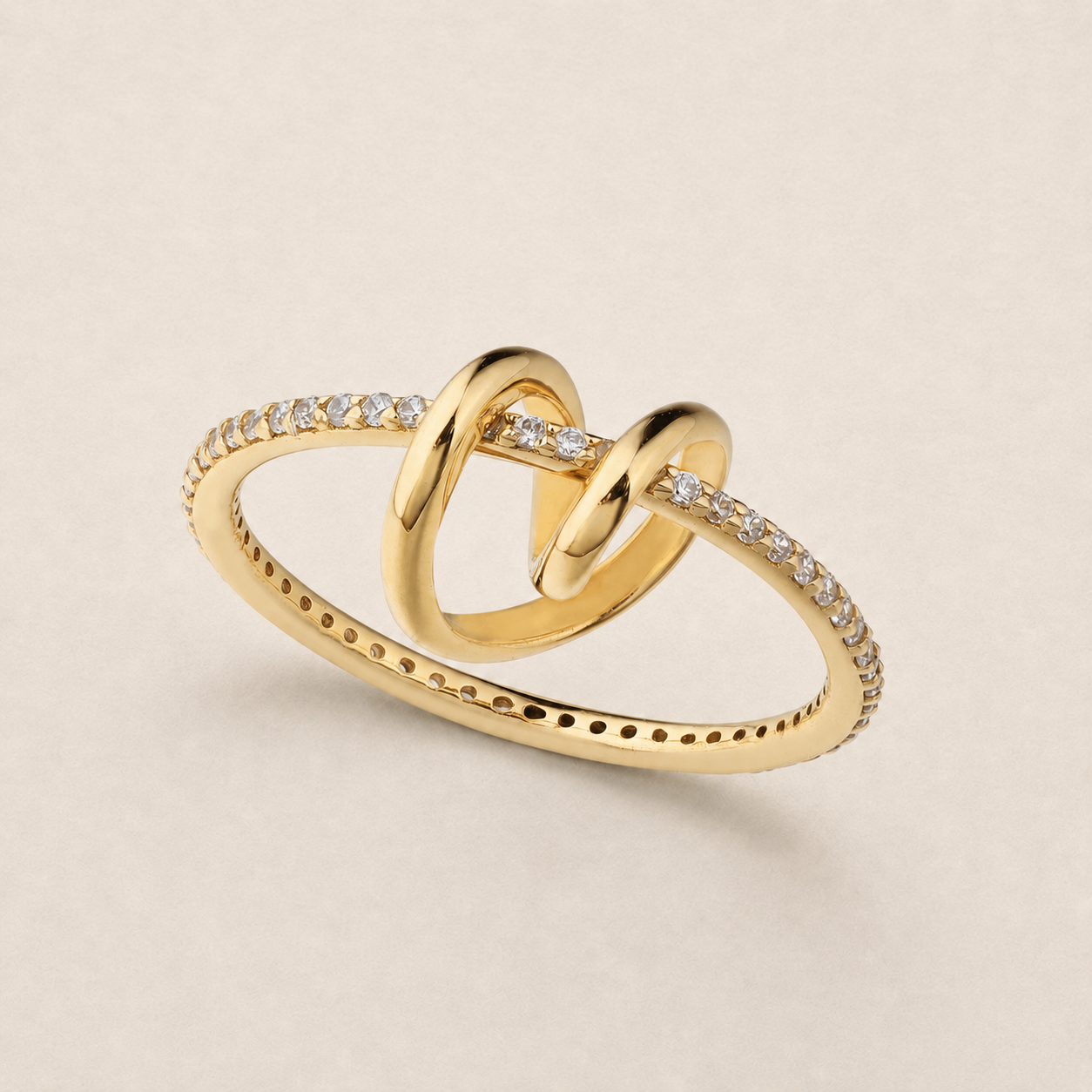 A gold ring featuring two interlocking knot-like loops, with pavé-set clear stones along the band and a row of small perforations on the lower band, photographed on a light beige background.