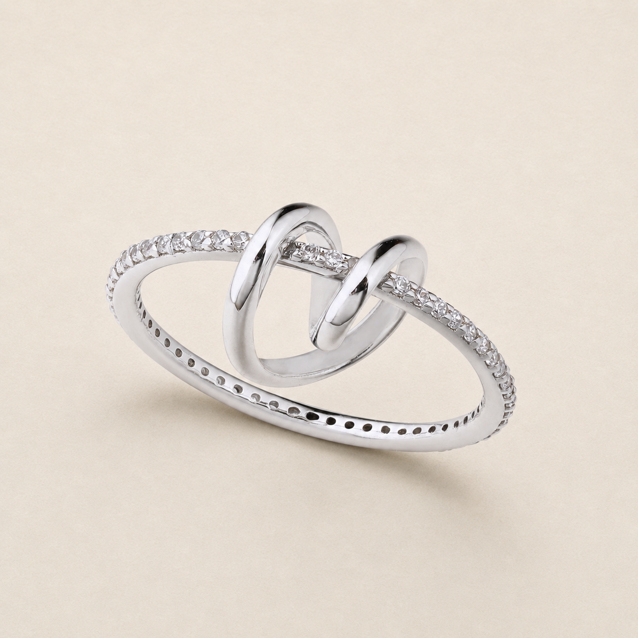 A silver ring featuring two interlocking heart-shaped loops, with pavé-set clear stones along the outer band, photographed on a light beige background.