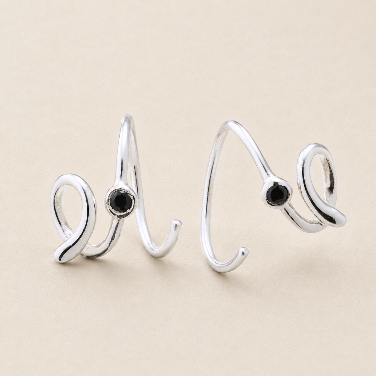 A pair of silver-toned open wrap rings with a small round black gemstone set into each band, displayed on a neutral beige background.