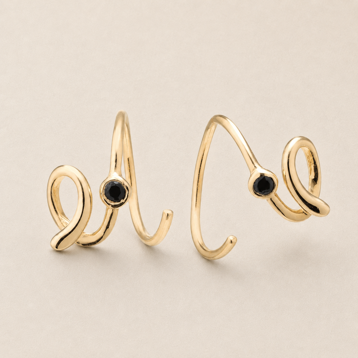 A pair of gold-toned open wrap rings with a small round black gemstone set into each band, displayed on a neutral beige background.