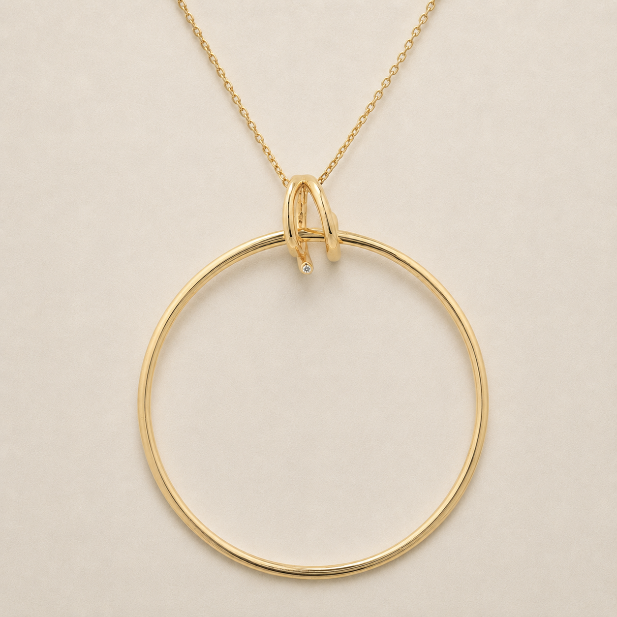 A gold necklace featuring a large, minimalist open circular pendant with a small, delicate interlocking loop at the top and a tiny clear gemstone accent, shown on a plain light beige background.