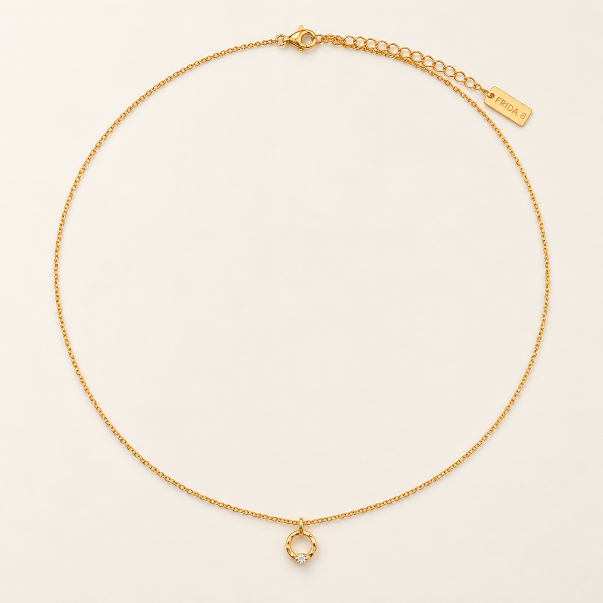 A delicate gold chain necklace with a small circular ring pendant set with tiny clear stones, displayed on a warm off-white background. A gold tag on the chain reads 'PRADA'.