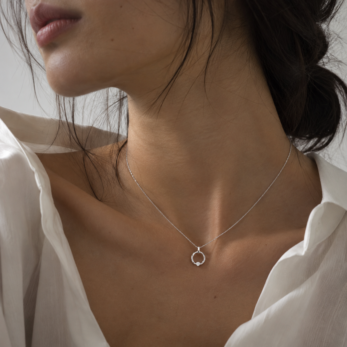 A close-up of a woman wearing a delicate silver necklace with a small open circular pendant, styled with a white blouse.