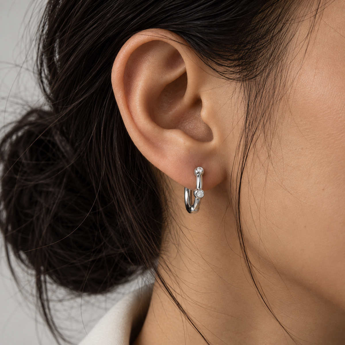 A close-up of a model's ear wearing a small silver hoop earring with a tiny clear gemstone accent.