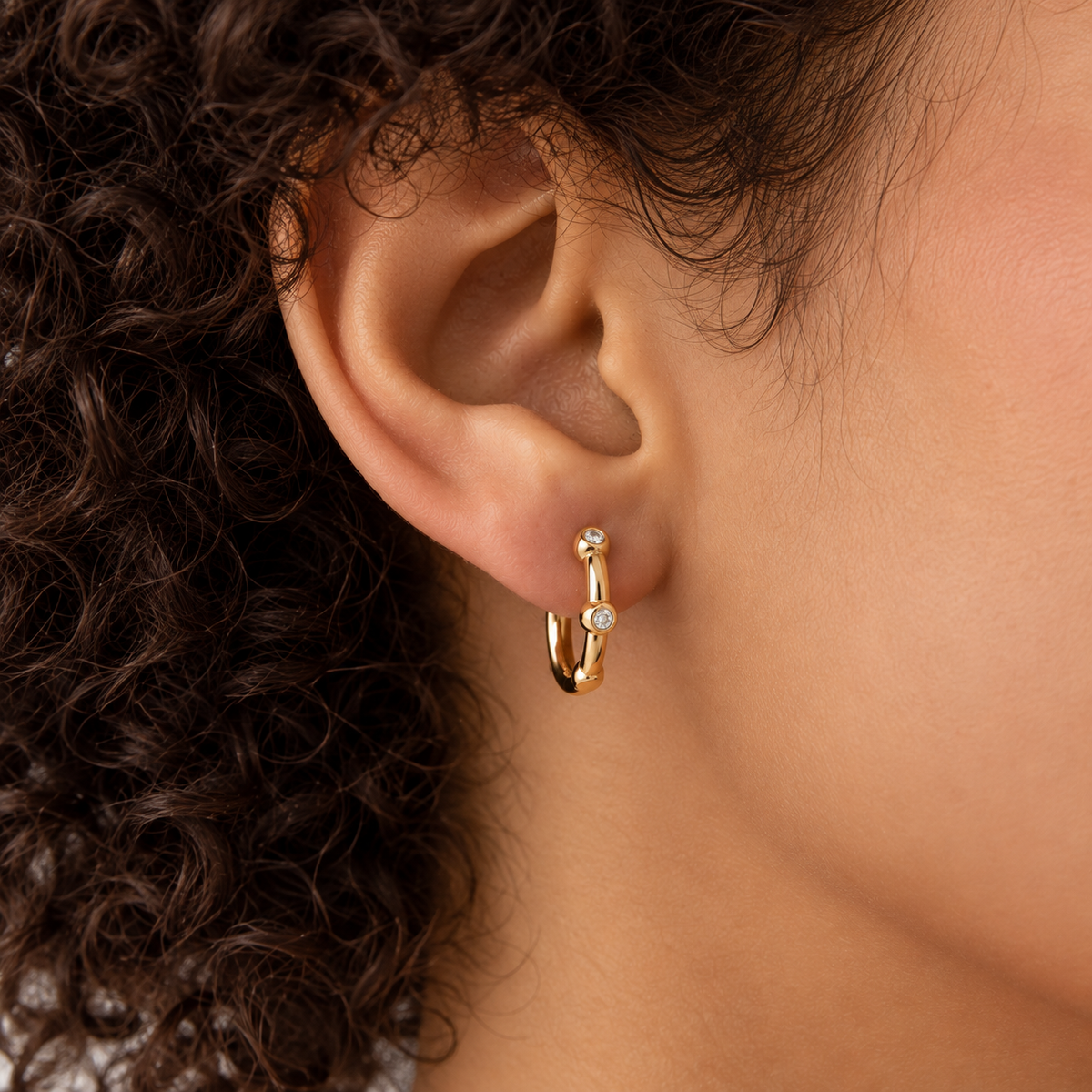 A close-up of a person's ear wearing a small gold huggie hoop earring with a tiny clear gemstone at the top and another small stone near the bottom.
