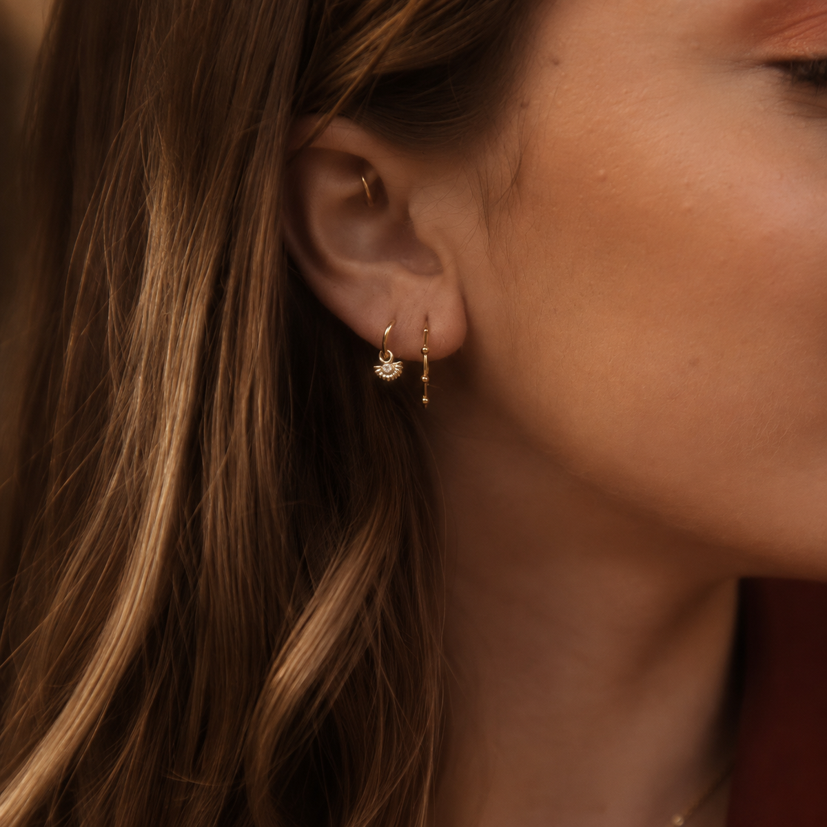 A close-up of a woman's ear wearing a pair of delicate gold huggie hoop earrings with small dangling charms, shown against her hair and skin.