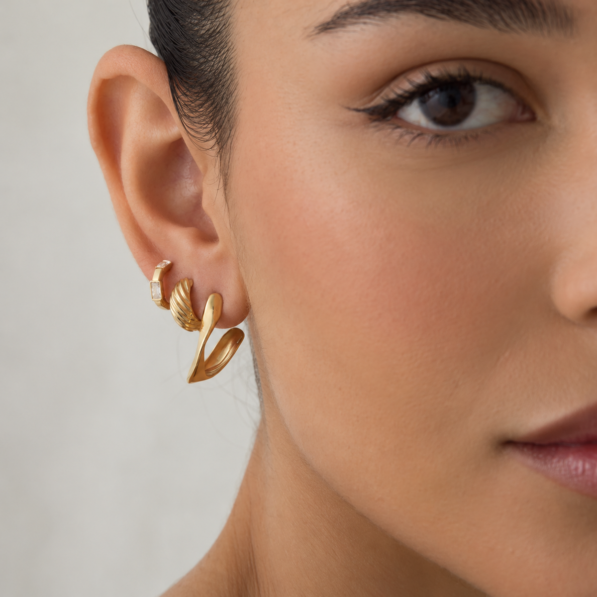 A close-up of a model's ear wearing a pair of gold huggie hoop earrings with twisted leaf-like dangling charms, set against a soft neutral background.