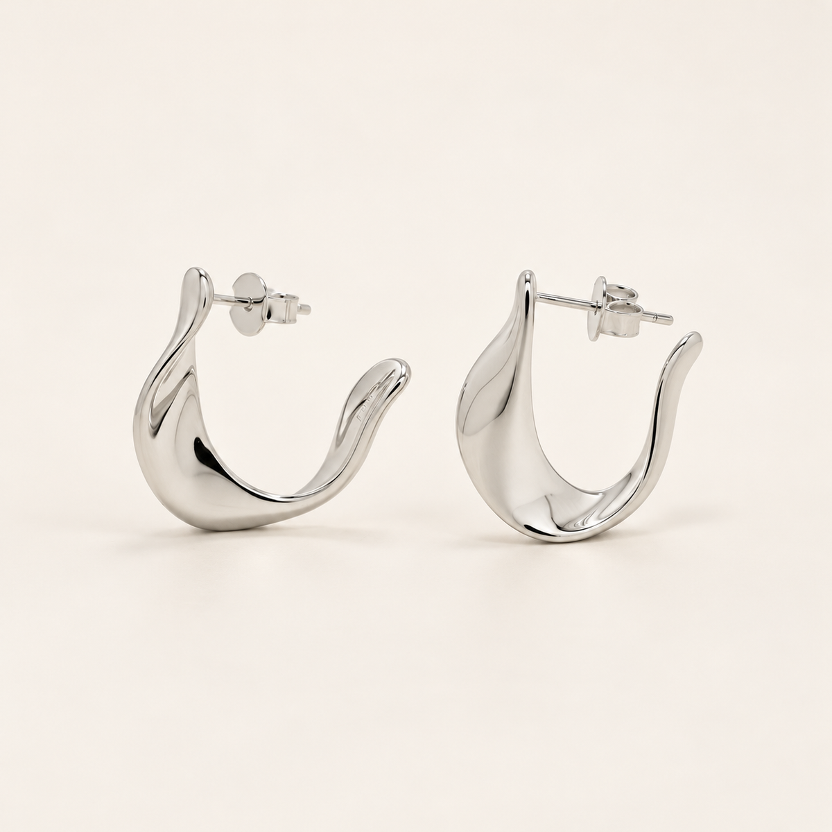A pair of polished silver huggie hoop earrings with a wavy, organic curved design, displayed on a clean, light beige background.