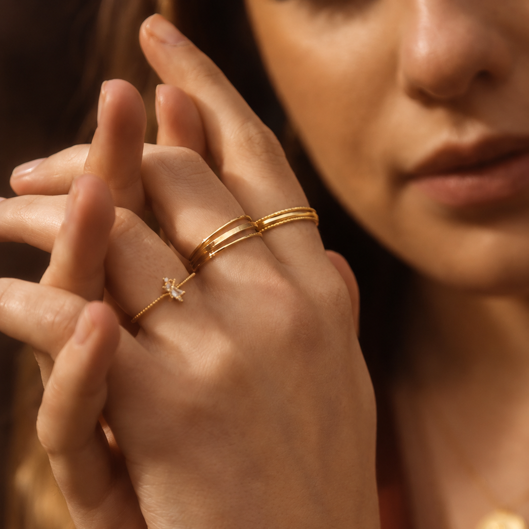 A close-up lifestyle shot of a model's hands wearing delicate gold rings, including a thin band with a small white crystal star and two thin bands with a small white crystal moon, against a warm, dark background.