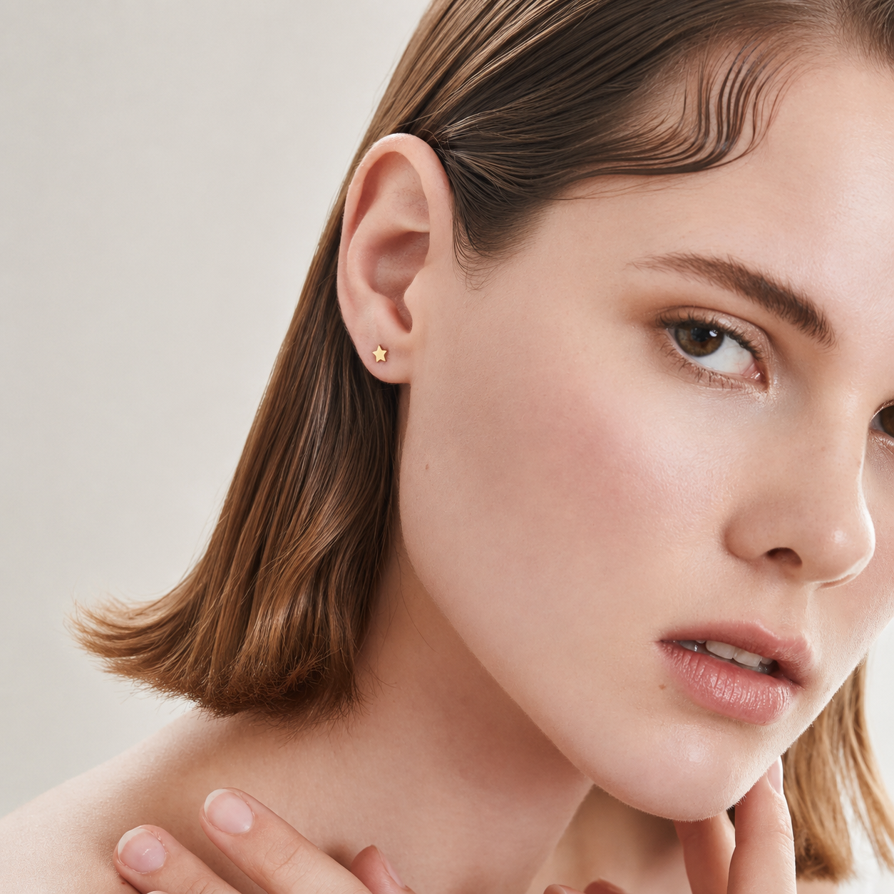 A close-up portrait of a woman with brown shoulder-length hair wearing a small gold stud earring, photographed against a soft light beige background.