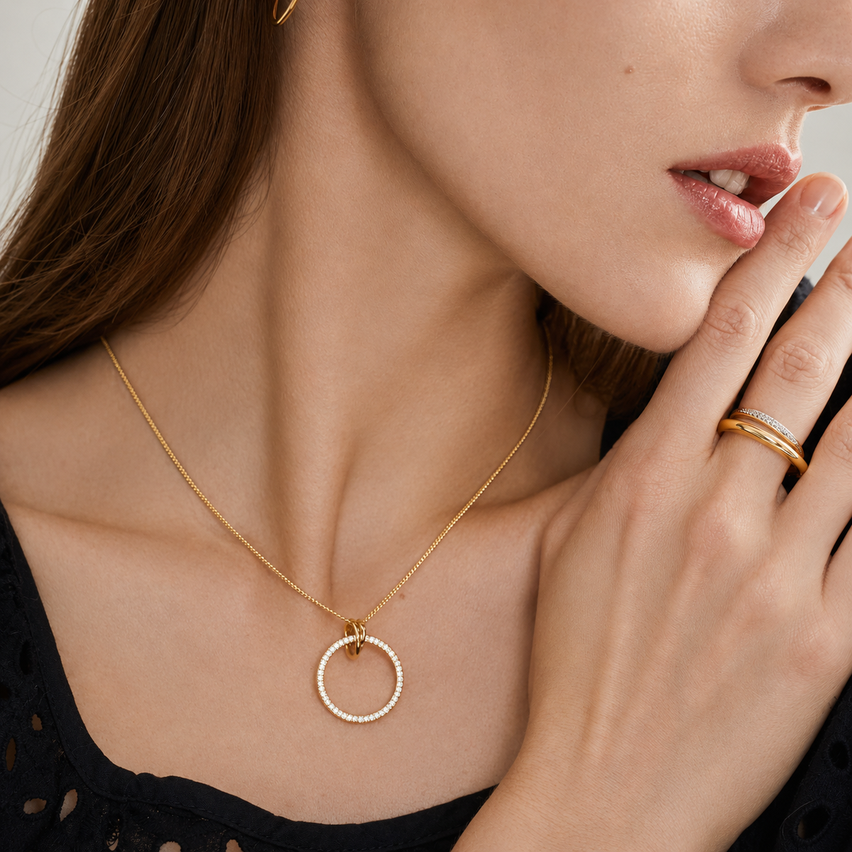 A close-up lifestyle shot of a model wearing a delicate gold chain necklace with a textured, bezel-set circular pendant. The model is also wearing gold rings on her fingers.