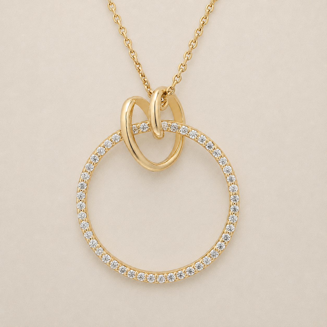 A gold pendant necklace featuring a large open circular ring paved with small clear stones, with two interlocking loop elements at the top and a delicate gold chain, shown on a light beige background.