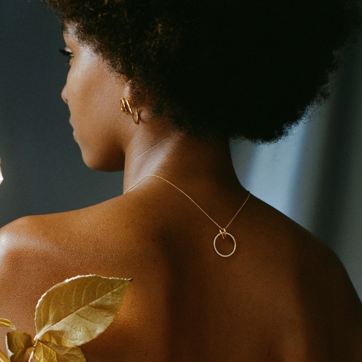 A woman shown from the back wearing a delicate gold chain necklace with a minimalist circular pendant, styled with a gold earring and a gold leaf accessory.