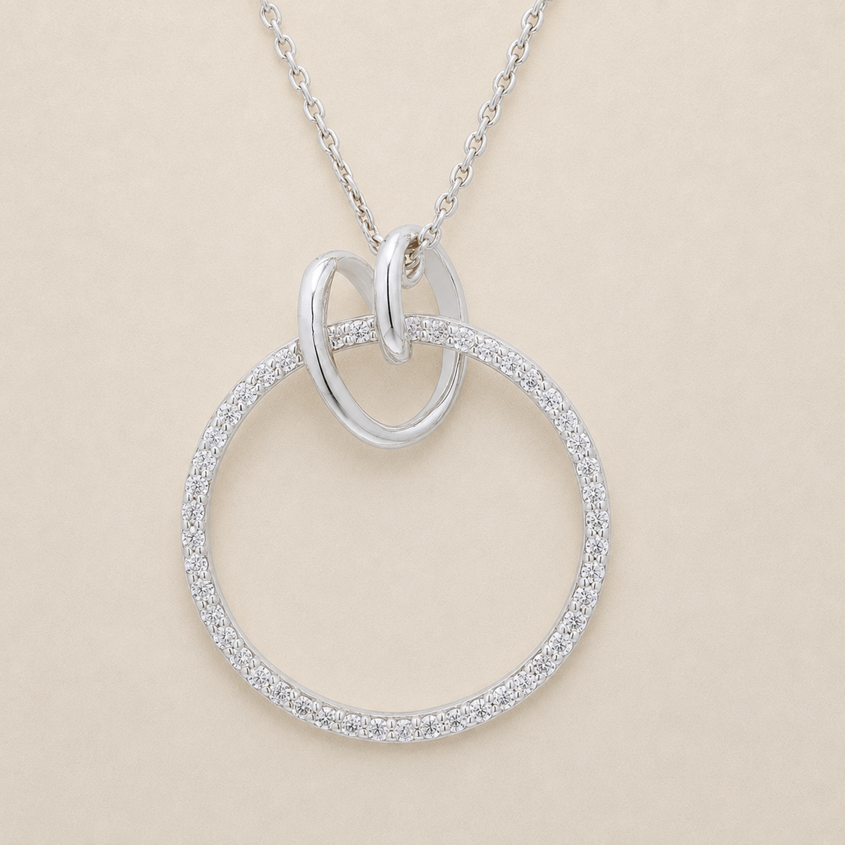 A silver necklace featuring a large open circle pendant paved with small clear stones, with a smaller polished oval loop at the top and a delicate chain, shown on a light beige background.