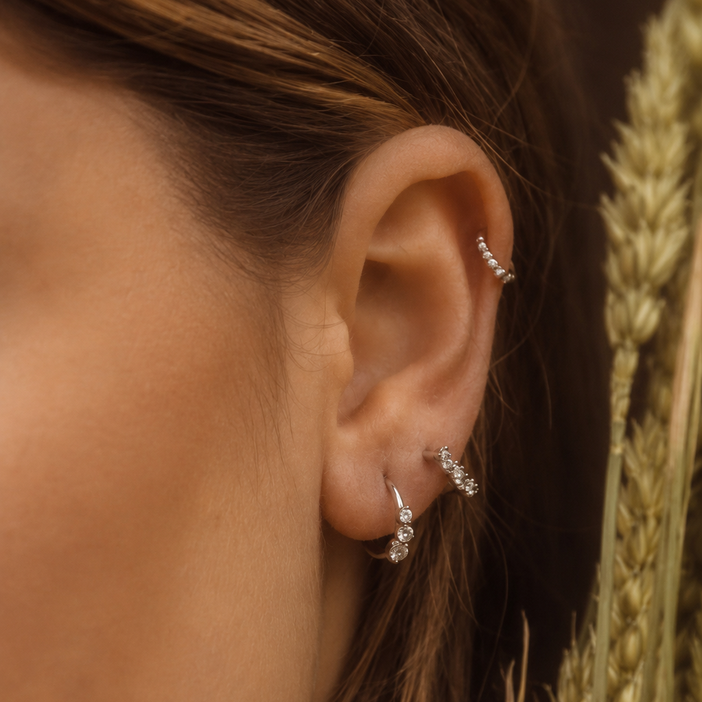 A close-up of a person's ear adorned with multiple delicate silver hoop and cartilage earrings, styled with golden wheat stalks against a warm brown background.