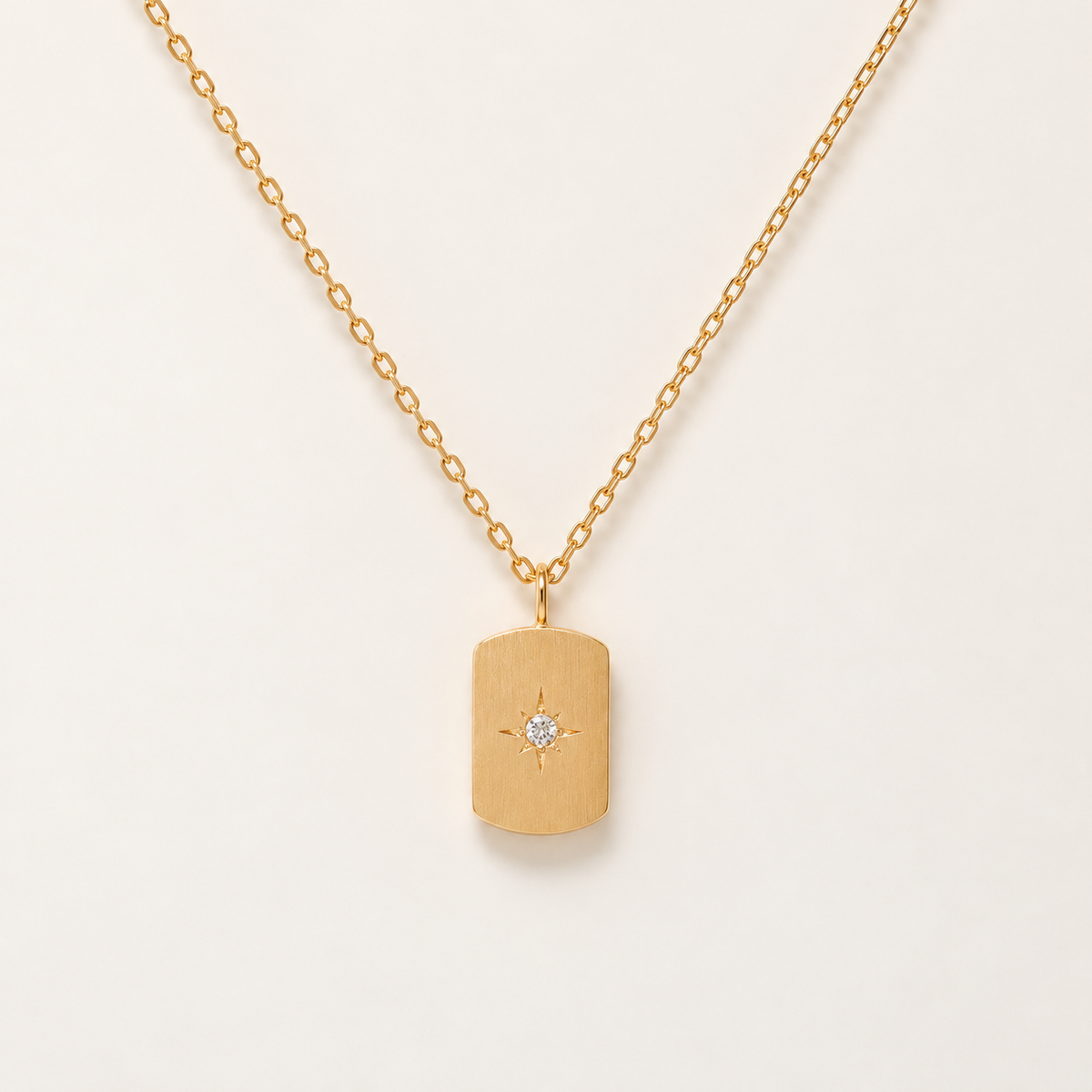 A gold chain necklace with a rectangular dog tag pendant featuring a small starburst detail and a clear central gemstone, shown on a light cream background.