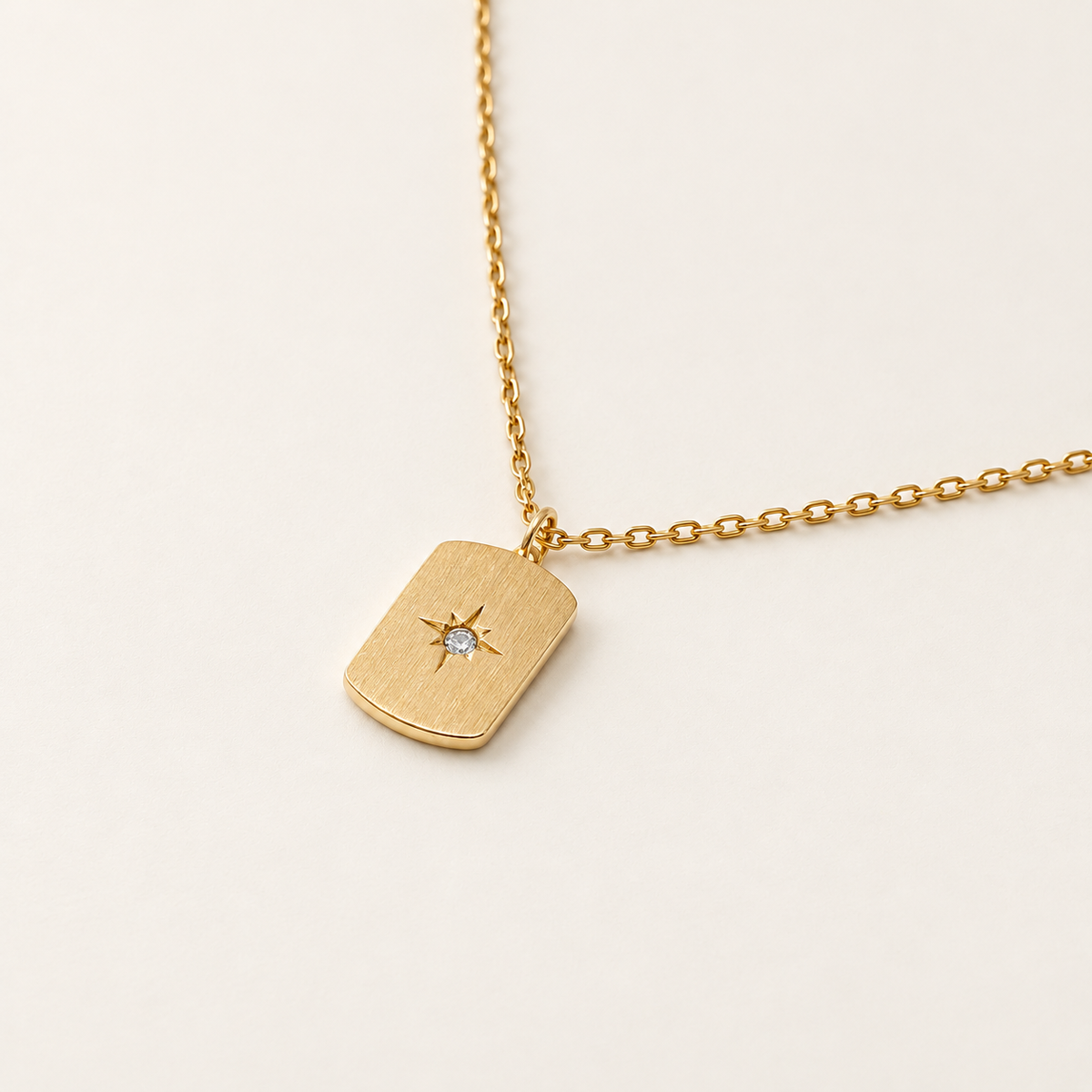 A gold dog tag necklace with a small starburst design and a clear central gemstone, shown on a delicate gold chain against a light cream background.