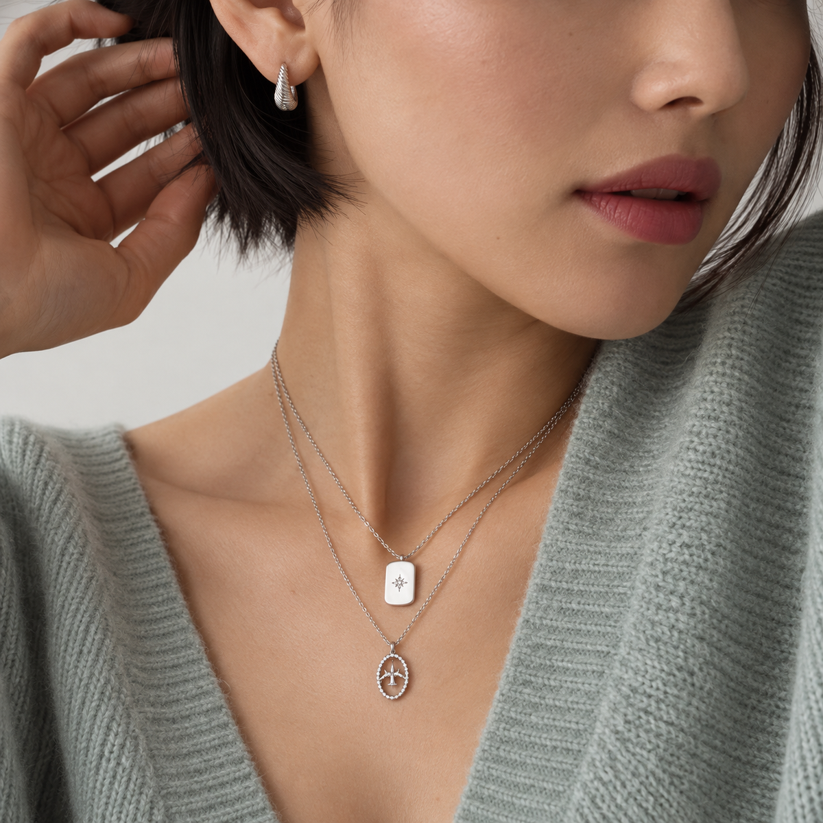 A close-up lifestyle shot of a model wearing a double-layered silver necklace with a square pendant and an oval peace-sign pendant, paired with a silver teardrop earring and a light green knit cardigan.