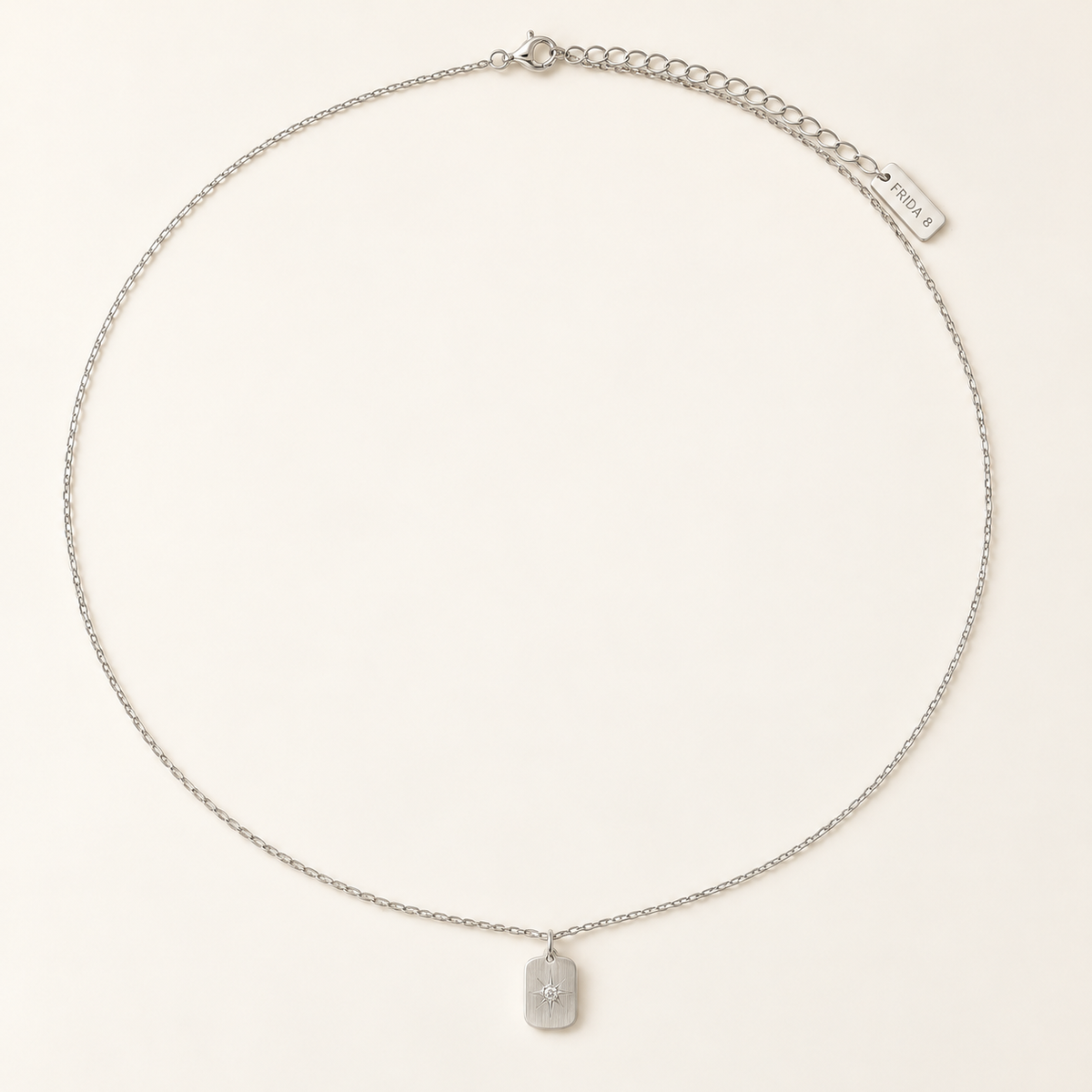 A delicate silver chain necklace with a small rectangular pendant featuring a raised cross, displayed on a clean, light beige background. The chain includes an adjustable lobster clasp and an extension chain with a brand tag that reads 'PRAIRIE'.