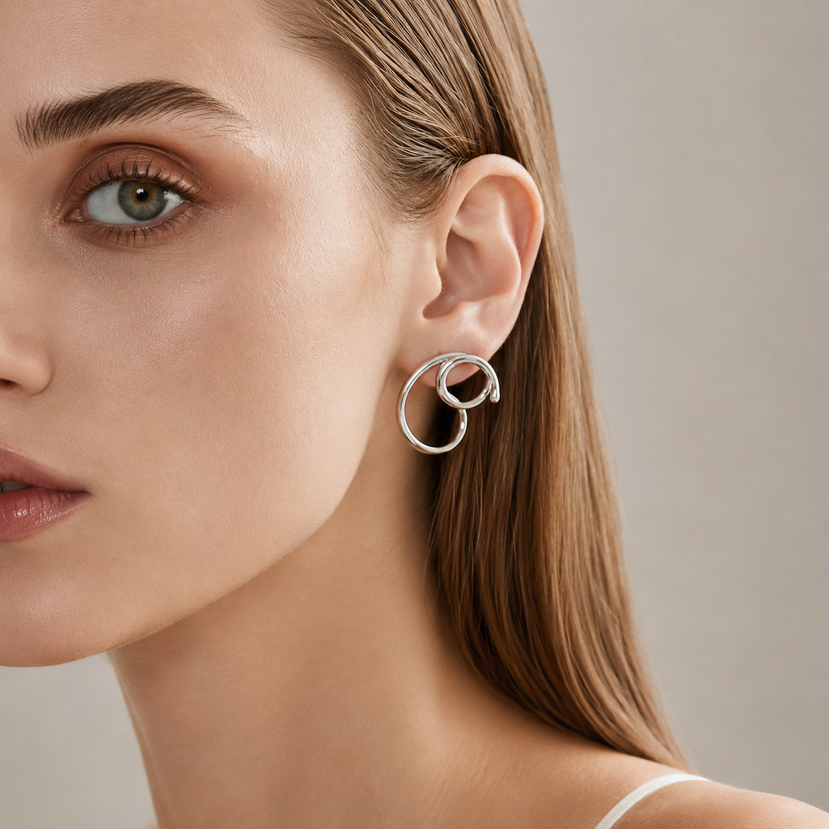 A close-up of a woman's ear wearing a polished silver double-ring hoop earring against a soft neutral background.