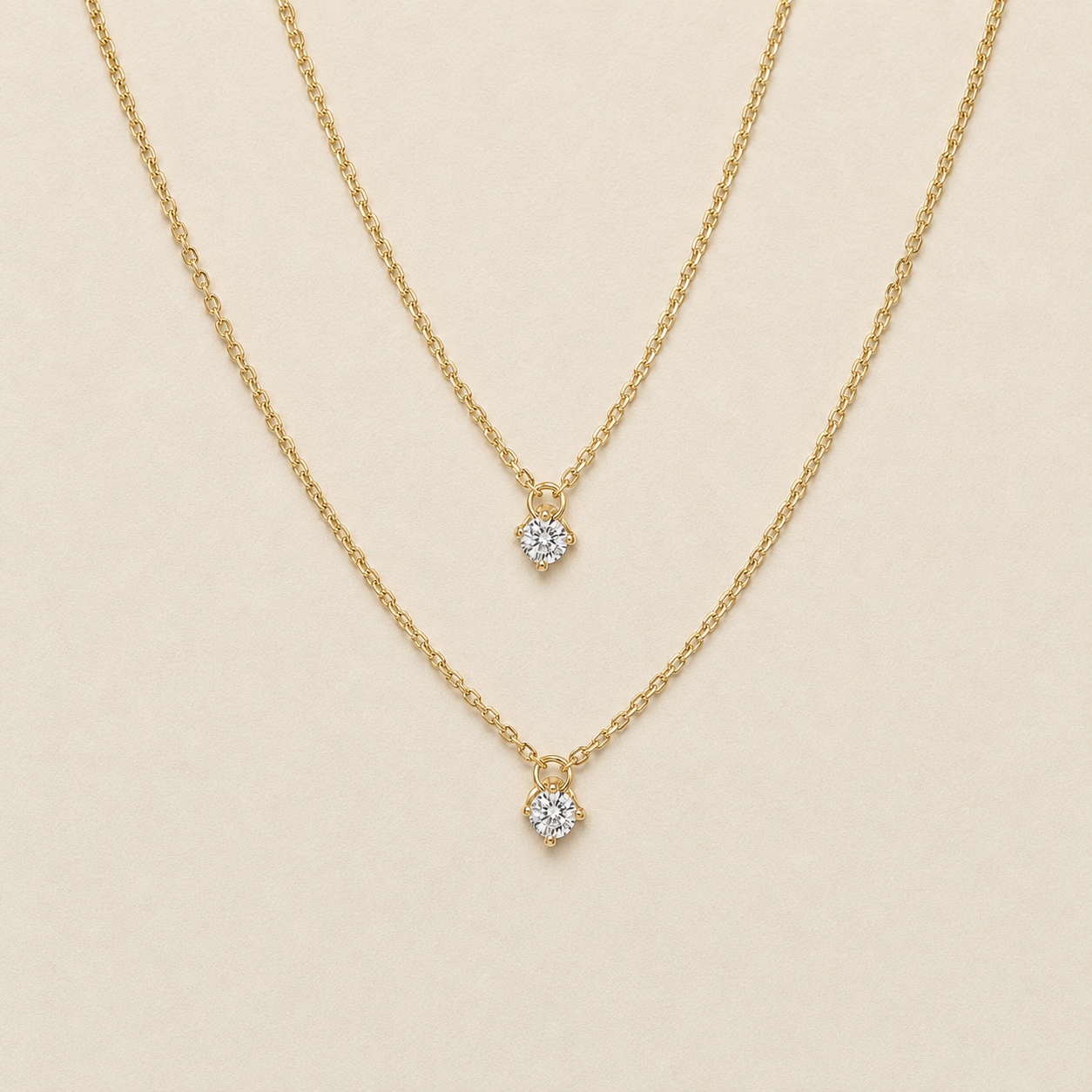 Two delicate gold chain necklaces with small round clear gemstone pendants, displayed on a light beige background.