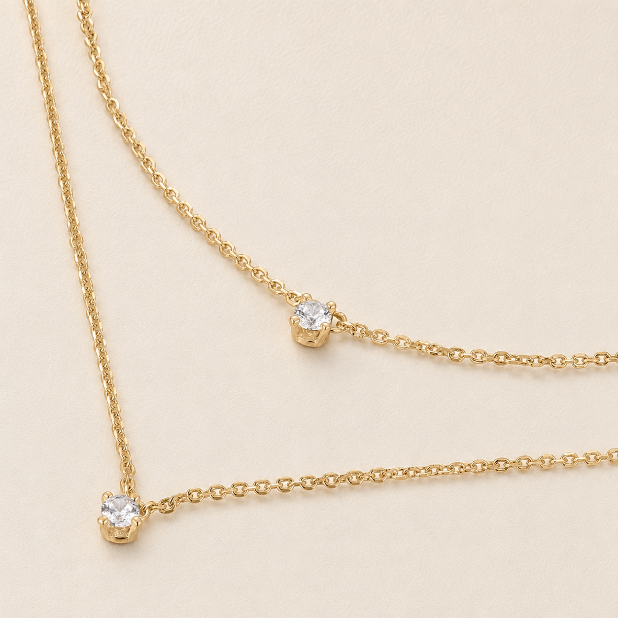Two delicate gold chain necklaces with single bezel-set clear gemstones, displayed on a light beige background.