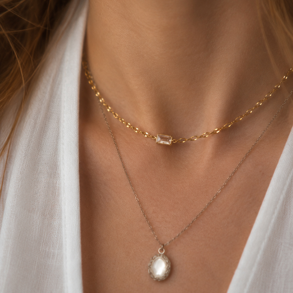 A close-up of a woman's neck and chest wearing two layered necklaces: a delicate gold chain choker with a small rectangular clear stone, and a longer silver-toned chain with a teardrop-shaped white pearl pendant.