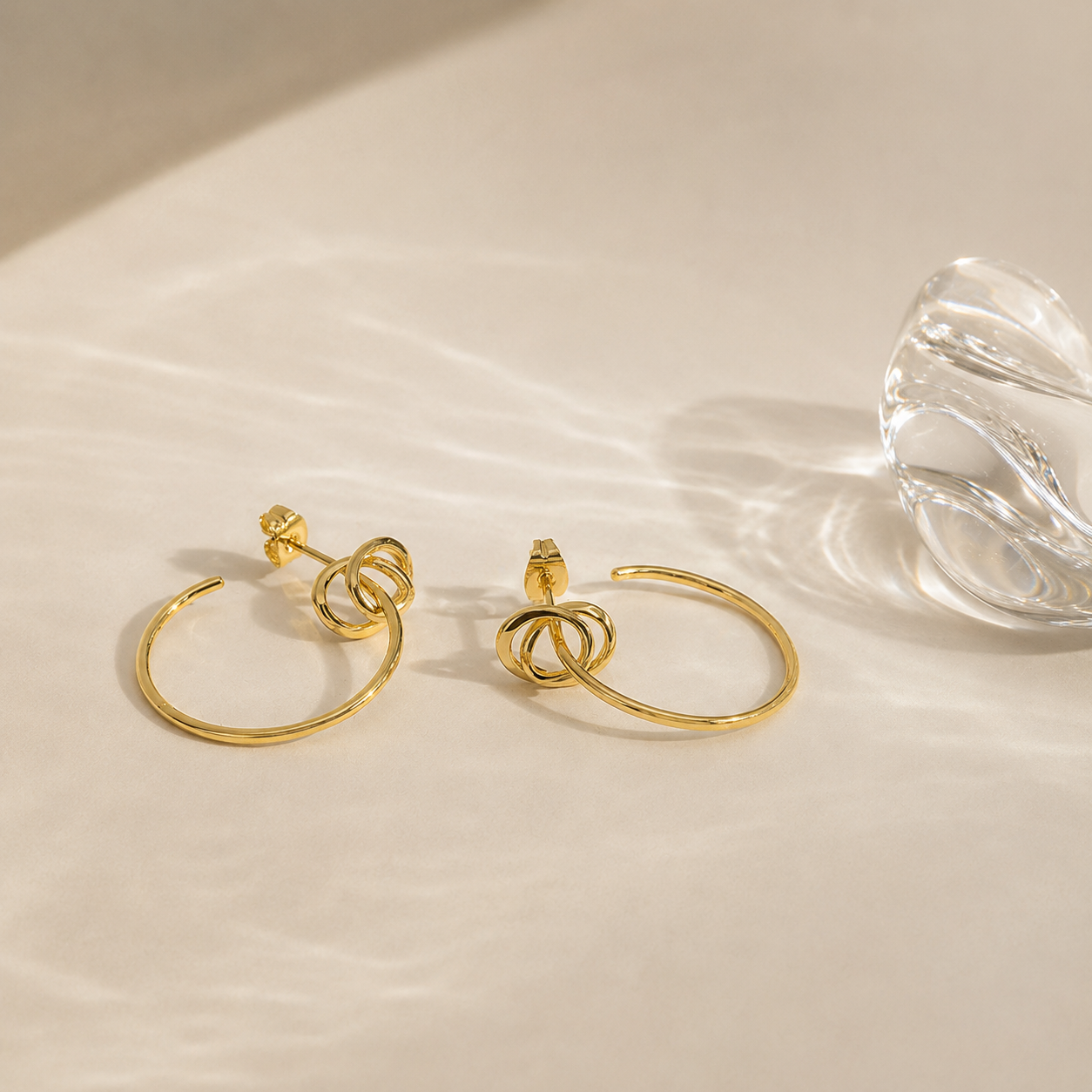 A pair of gold hoop earrings with a knotted loop design at the front, displayed on a warm beige surface next to a clear glass object.