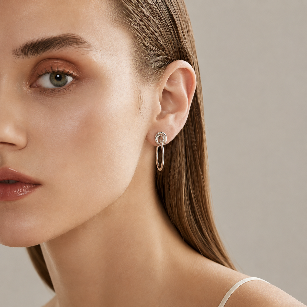 A close-up of a woman's ear wearing a delicate silver hoop earring with a small spiral detail on the post, set against a soft neutral background.
