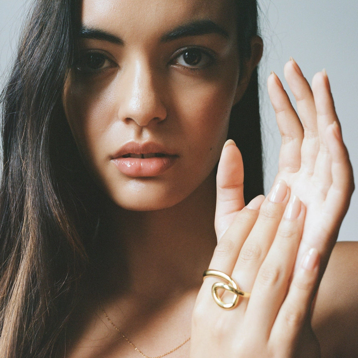 A close-up portrait of a woman with long dark hair wearing a delicate gold necklace and a gold ring on her hand, set against a soft gray studio background.