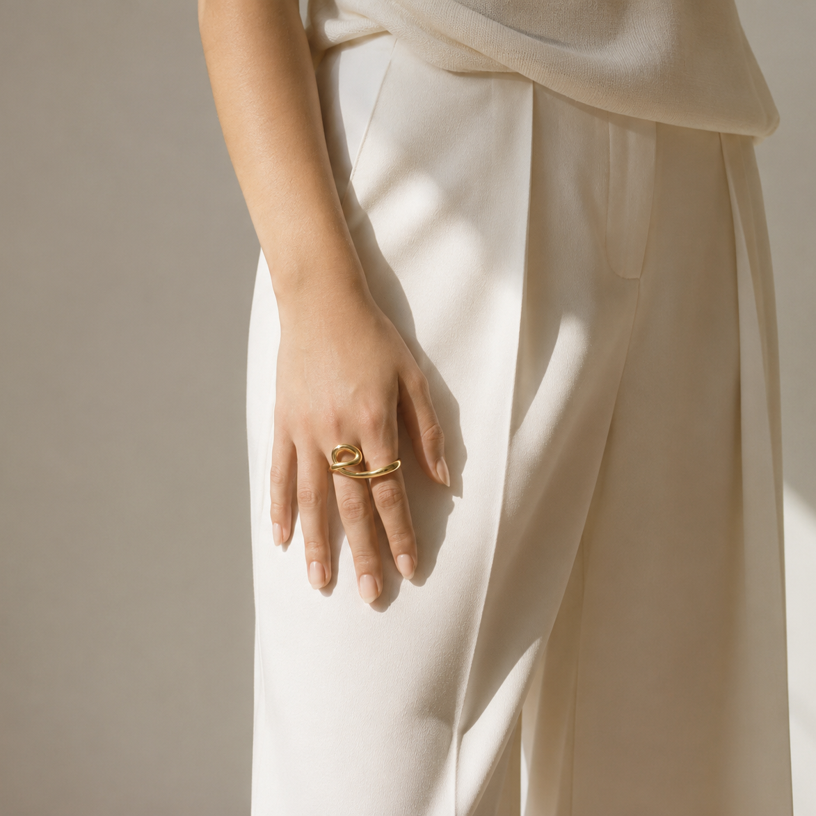 A close-up of a person wearing a cream-colored draped top and wide-leg trousers, with a gold ring on one finger, set against a warm beige background with soft shadows.