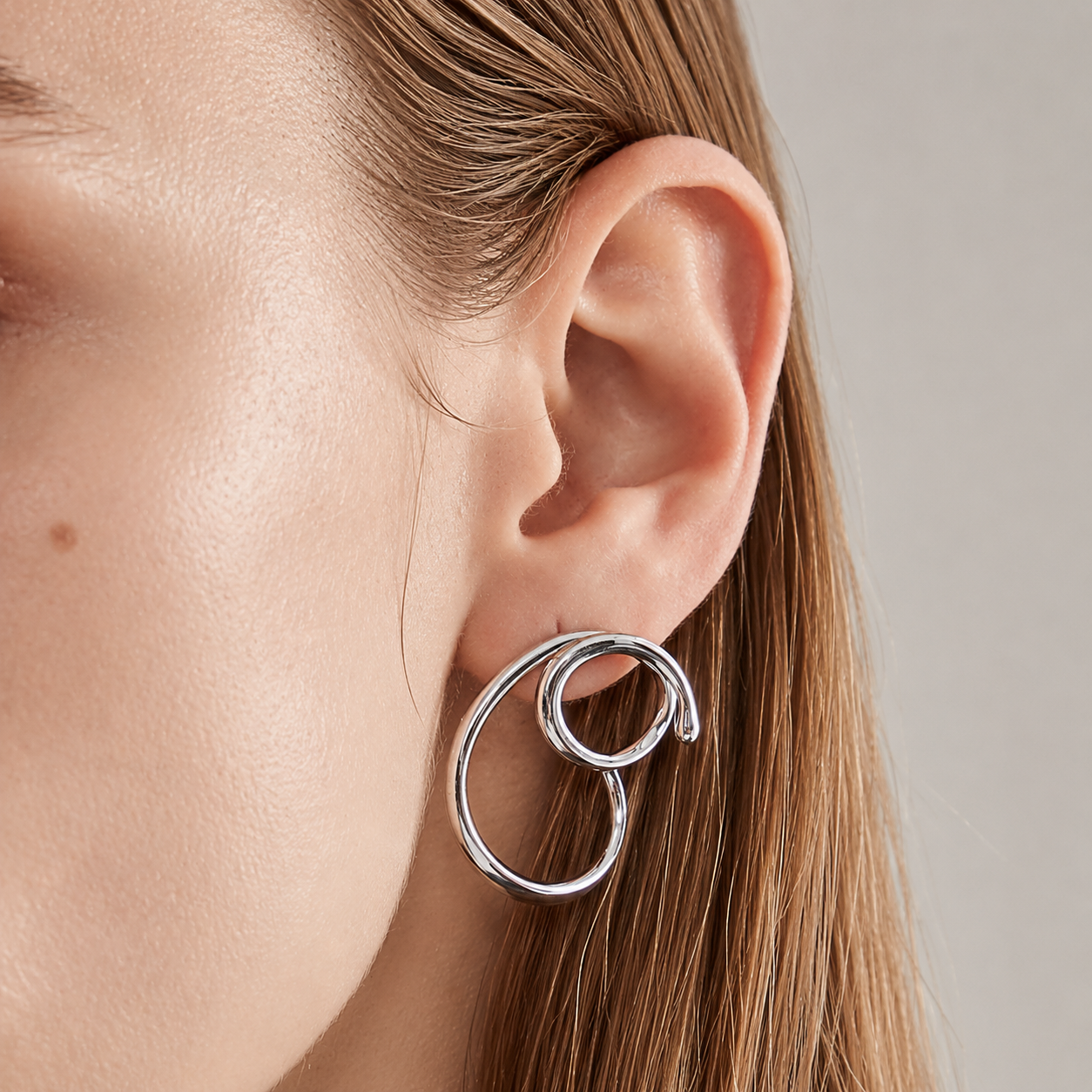 A close-up of a model's ear wearing a polished silver spiral hoop earring against a neutral light gray background.